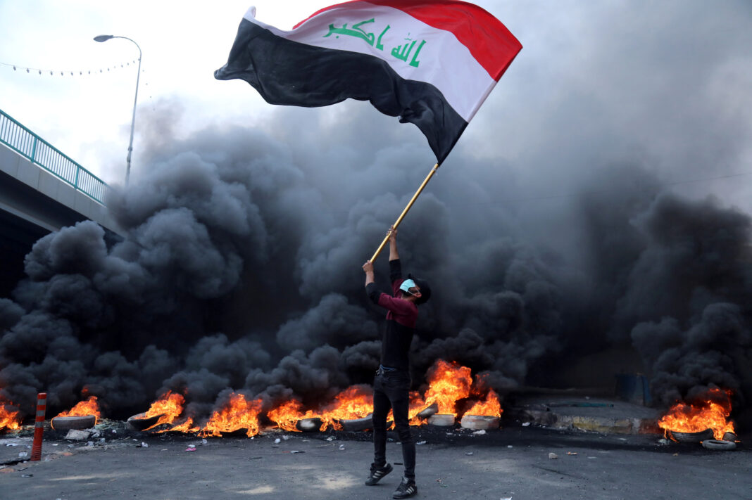 Protester holds an Iraqi flag next to burning tires during ongoing anti-government protests in Kerbala